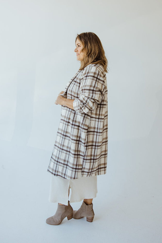 A person standing sideways wears the Andrea by Unit LIGHTWEIGHT PLAID MIDI BLAZER over a white dress and gray ankle boots, with hands clasped in front.