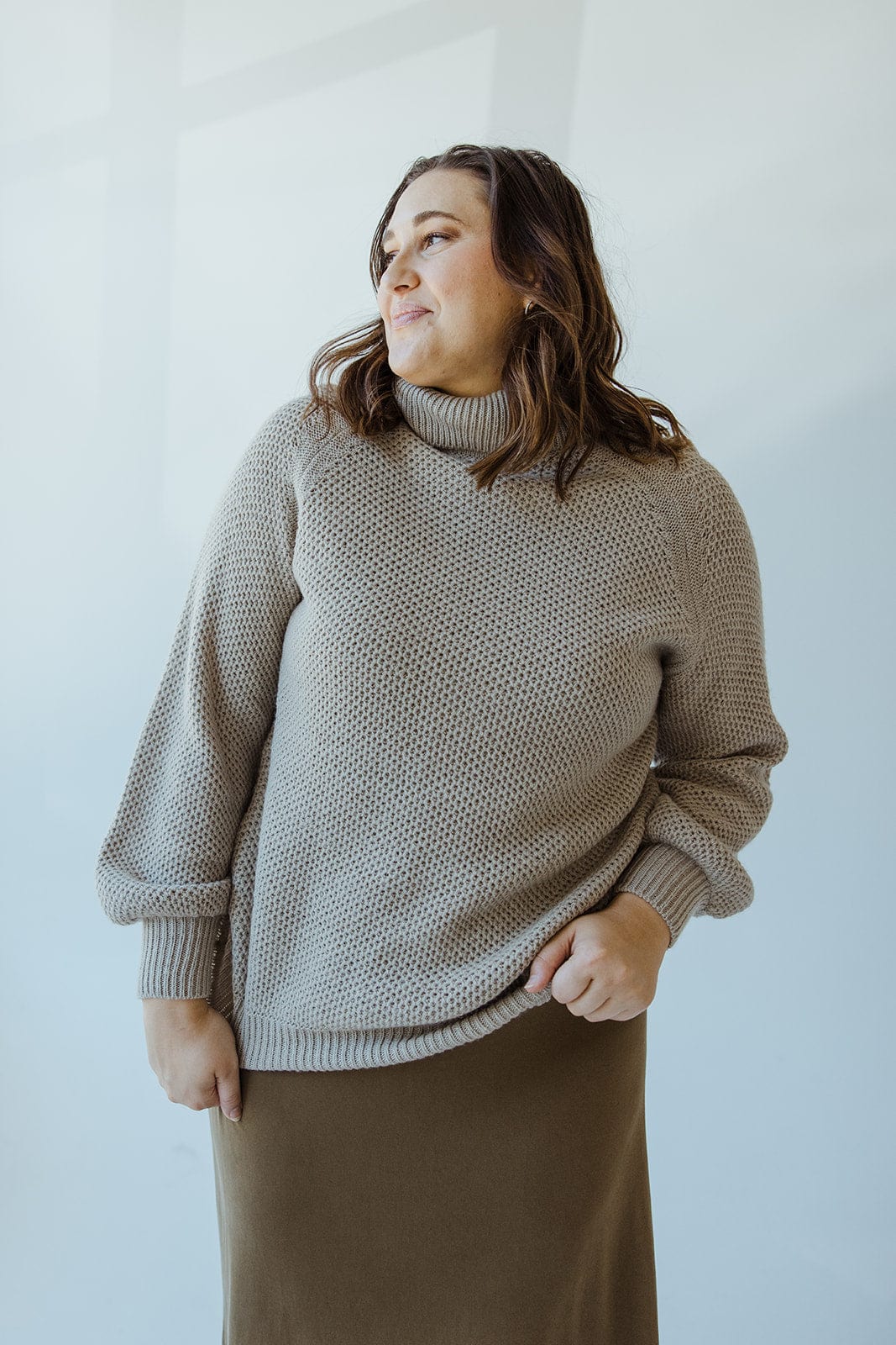 A person with shoulder-length hair is wearing a Yesta Lightweight Raglan Sleeve Turtleneck in Dried Sage and a long brown skirt, standing against a light-colored background.