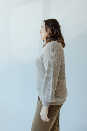 A person with shoulder-length hair is standing in profile against a light-colored background, wearing a Yesta textured beige lightweight raglan sleeve turtleneck in dried sage and brown pants.