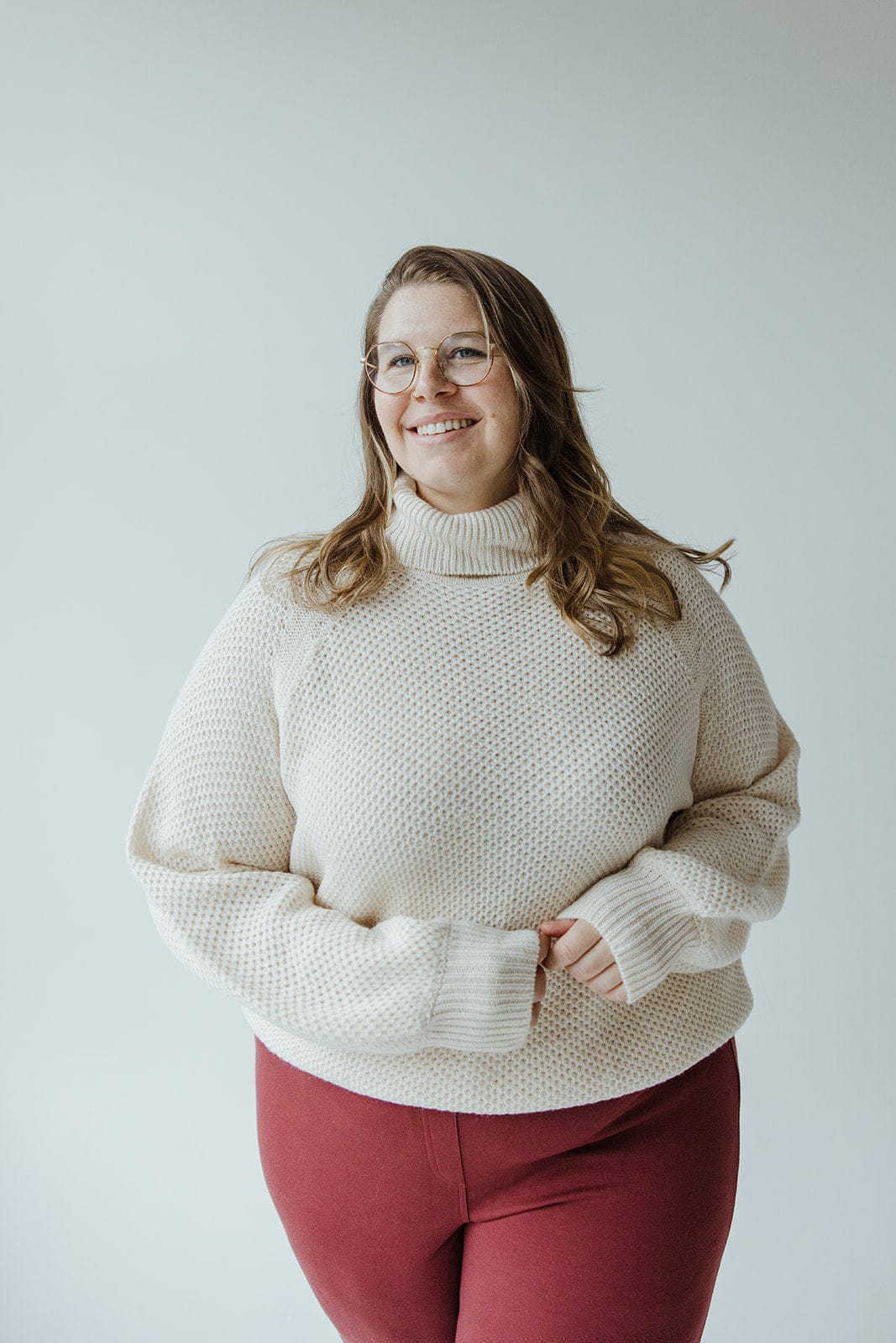 A person with long hair and glasses, dressed in a snowy white Yesta Lightweight Raglan Sleeve Turtleneck and red pants, stands in front of a plain background, smiling.