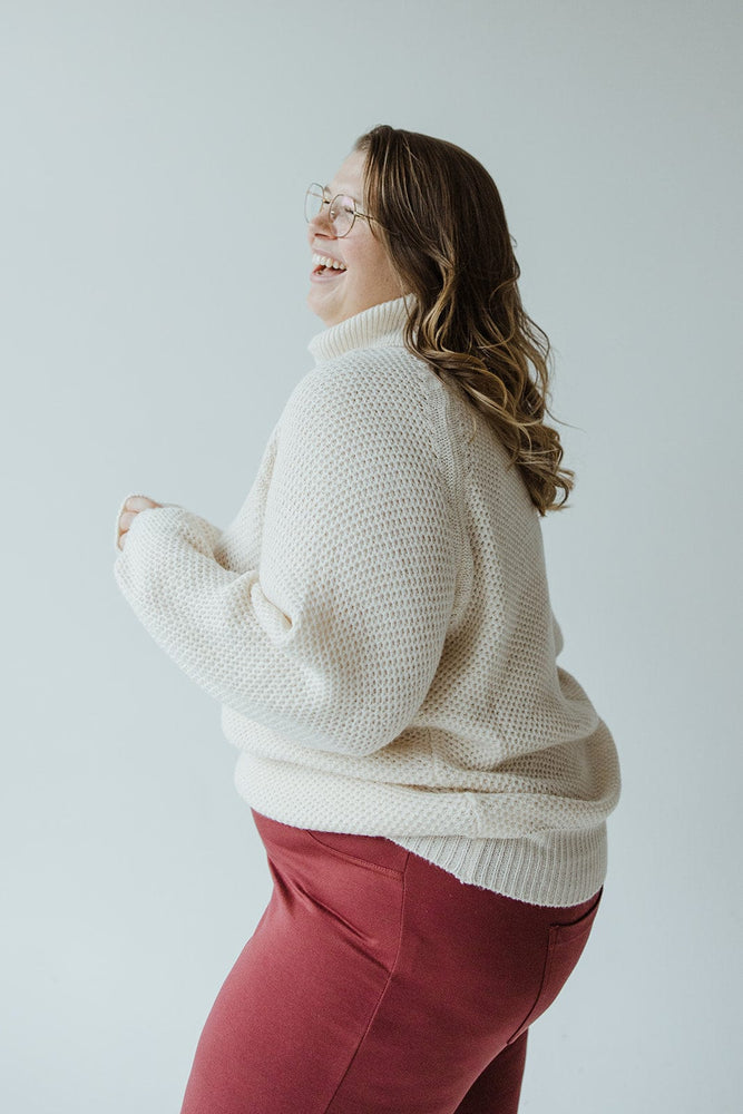 A person with long hair smiles and laughs while wearing glasses, the Lightweight Raglan Sleeve Turtleneck in Snowy White by Yesta, and red pants, standing against a plain background.