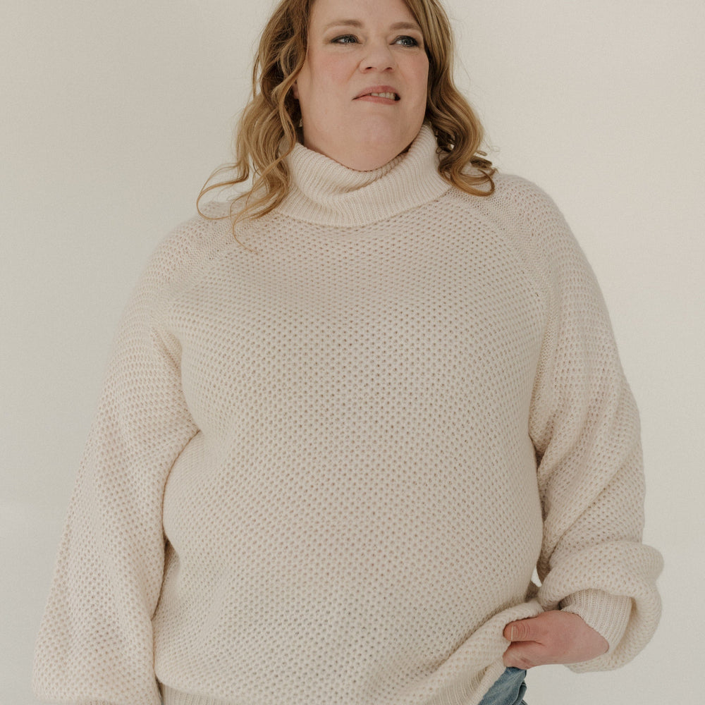 Person wearing a beige sweater and blue jeans against a plain background