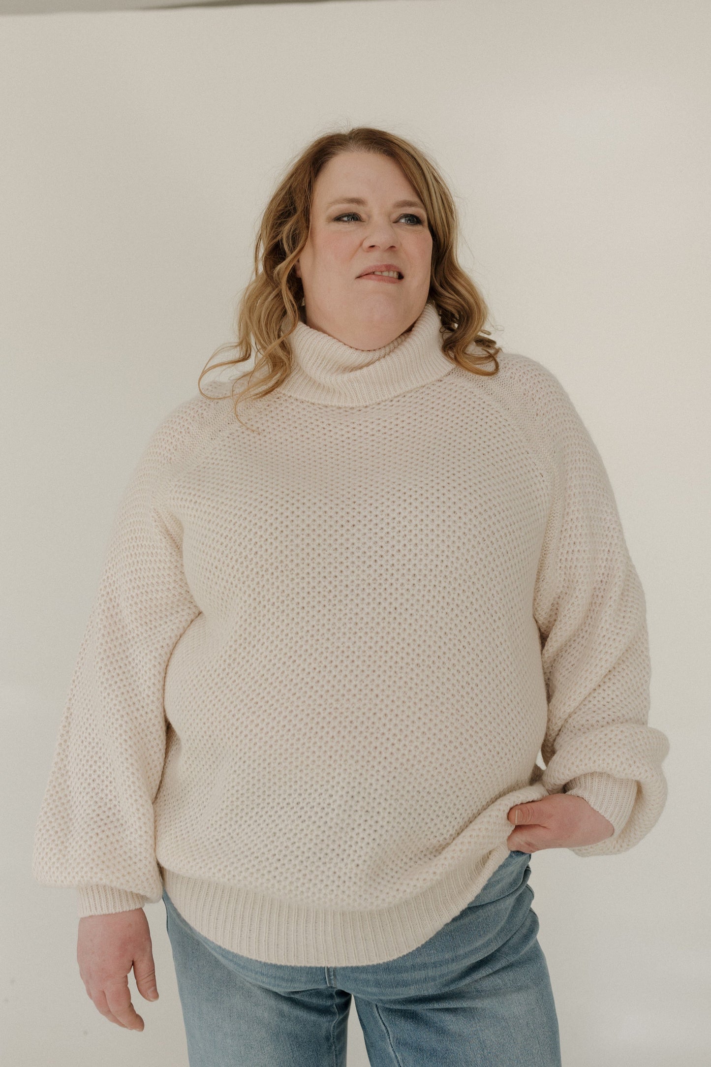 Person wearing a beige sweater and blue jeans against a plain background