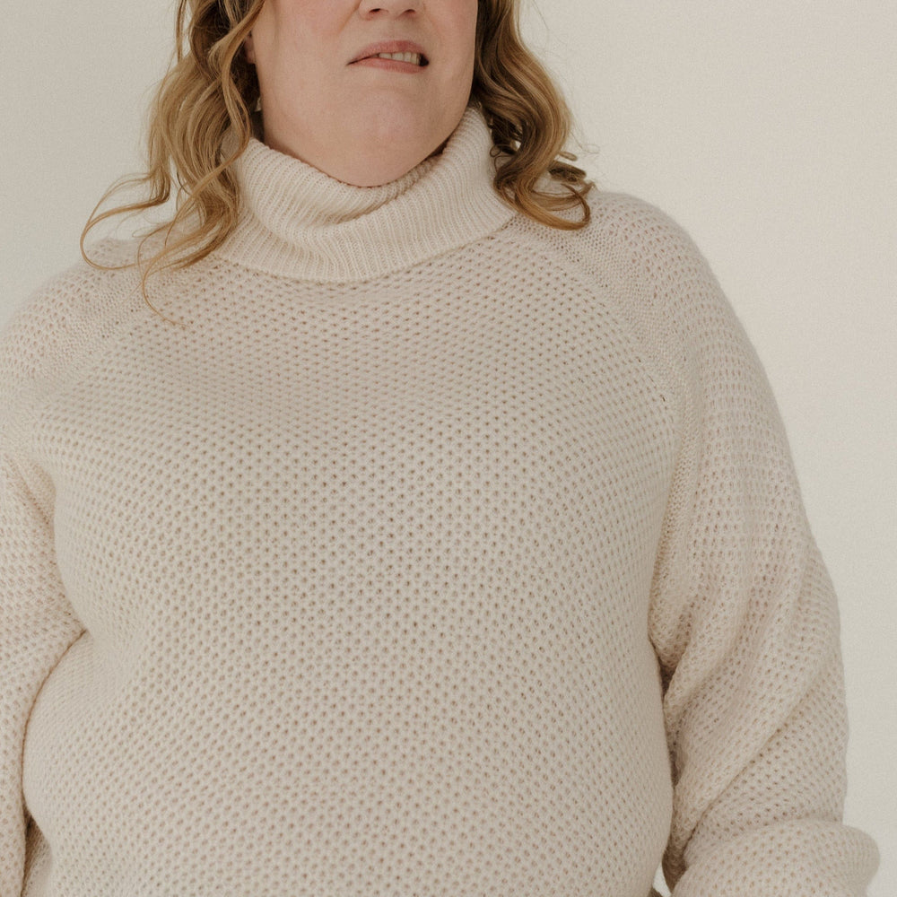 Person wearing a beige sweater and blue jeans against a plain background