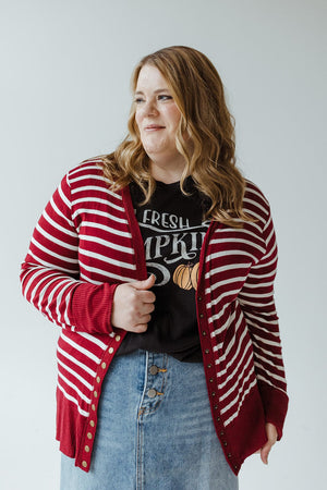 A woman with long hair is wearing a Zenana Lightweight Striped Snap Cardigan in Cranberry and Ivory over a black t-shirt featuring a "Fresh Pumpkin" design and a denim skirt, smiling with a relaxed expression.