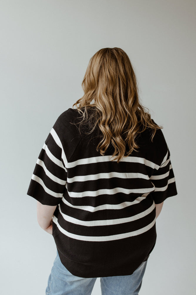 A person with long wavy hair stands with their back to the camera against a neutral background, dressed in Yesta's Lightweight Stripe Sweater in Black and Ecru paired with jeans. The ultra-flattering stripes beautifully accentuate the sweater's elegant round neckline.