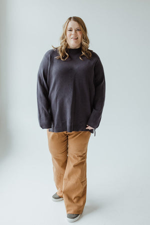 A person with long hair, wearing a dark blue-grey, lightweight Yesta sweater featuring subtle zipper details, light brown pants, and grey shoes stands against a plain white background.