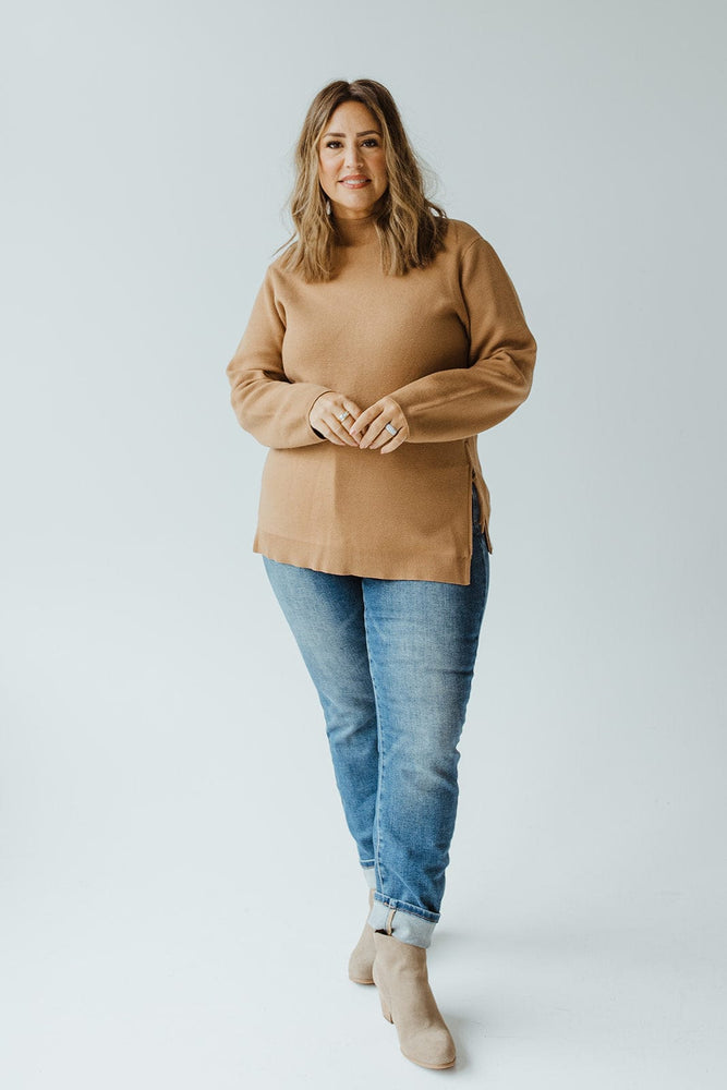 A woman with shoulder-length hair wearing a Yesta LIGHTWEIGHT SWEATER WITH ZIPPER DETAIL IN DARK CAMEL, blue jeans, and beige ankle boots stands against a plain white background.