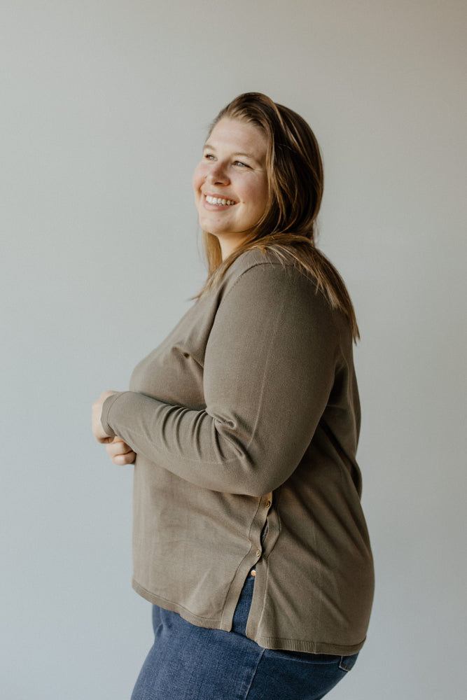 A person with long hair stands facing away, wearing a Yesta Lightweight V-Neck Sweater in Soft Army paired with blue jeans, set against a plain background.