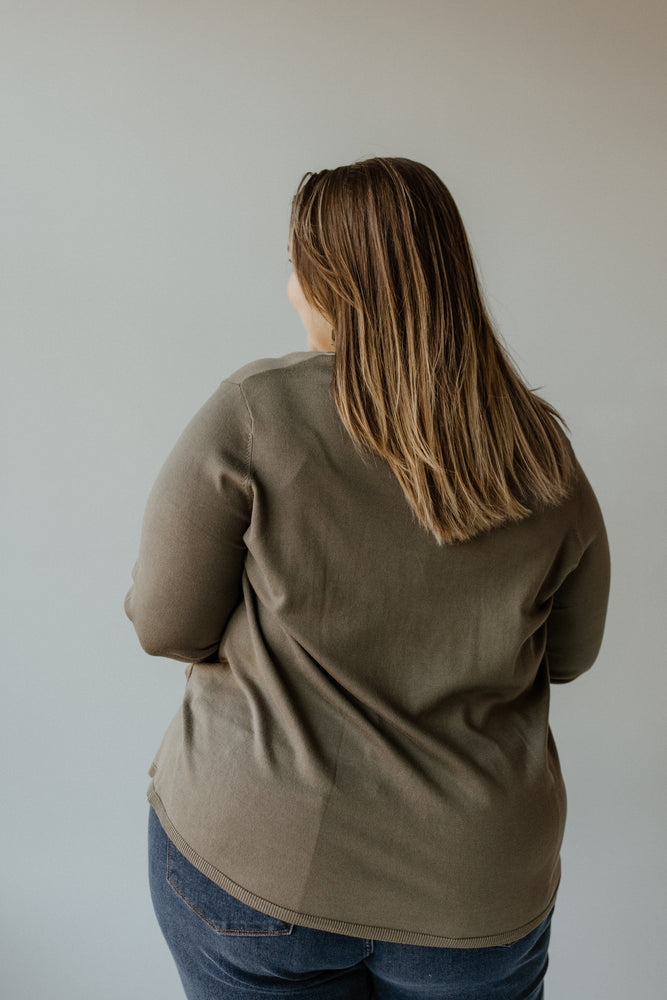 A person with long hair stands facing away, wearing a Yesta Lightweight V-Neck Sweater in Soft Army paired with blue jeans, set against a plain background.