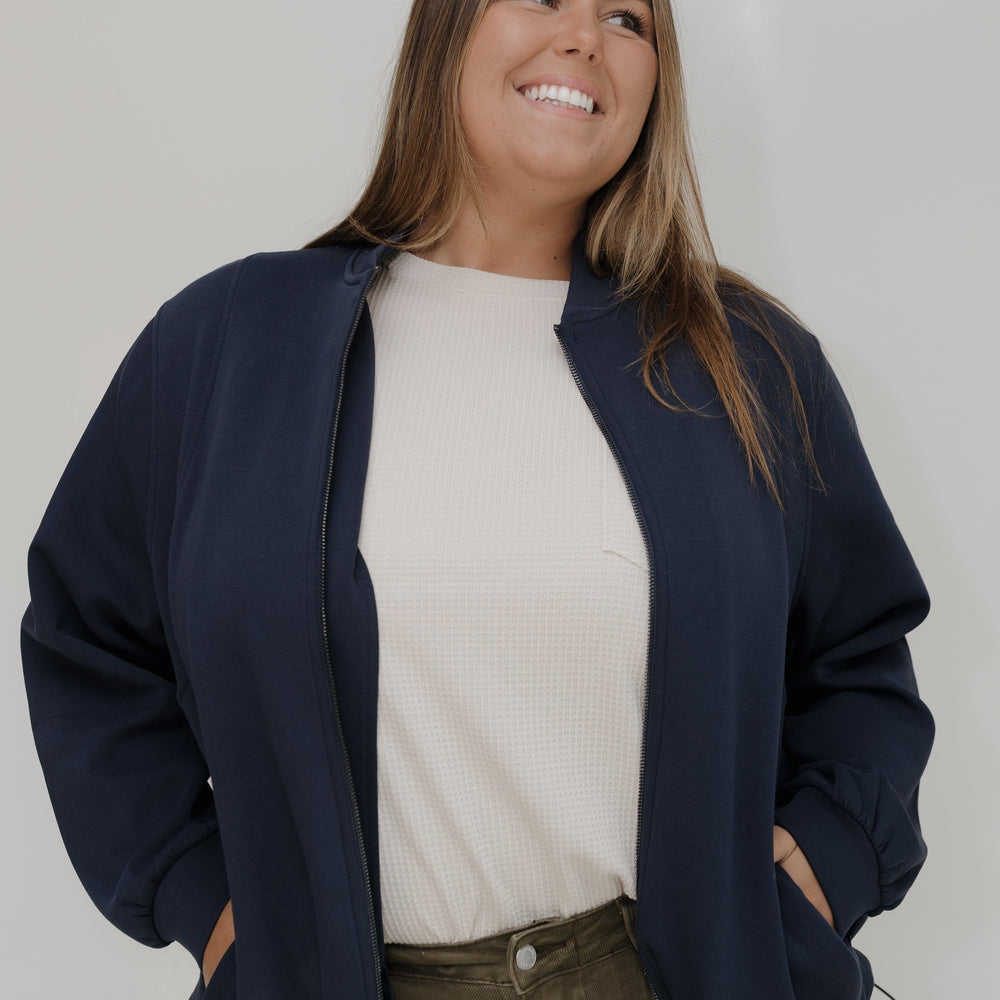 Woman wearing a navy blue jacket over a white shirt and brown pants against a plain background