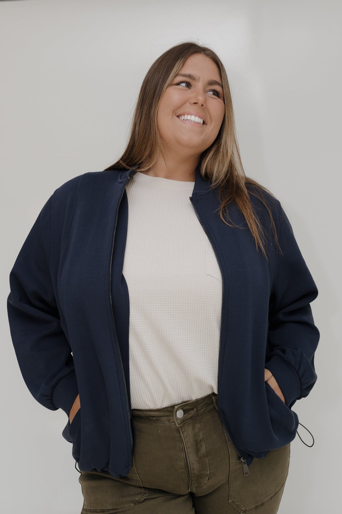 Woman wearing a navy blue jacket over a white shirt and brown pants against a plain background