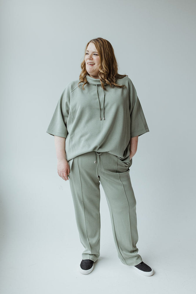 A woman with long hair is standing and smiling, showcasing a LIGHT AS AIR COWL NECK TEE IN ECHEVERRIA by Yesta with a comfortable oversized fit and matching light green pants, complemented by black slip-on shoes, all set against a plain light gray background.