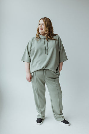 A woman with long hair is standing and smiling, showcasing a LIGHT AS AIR COWL NECK TEE IN ECHEVERRIA by Yesta with a comfortable oversized fit and matching light green pants, complemented by black slip-on shoes, all set against a plain light gray background.