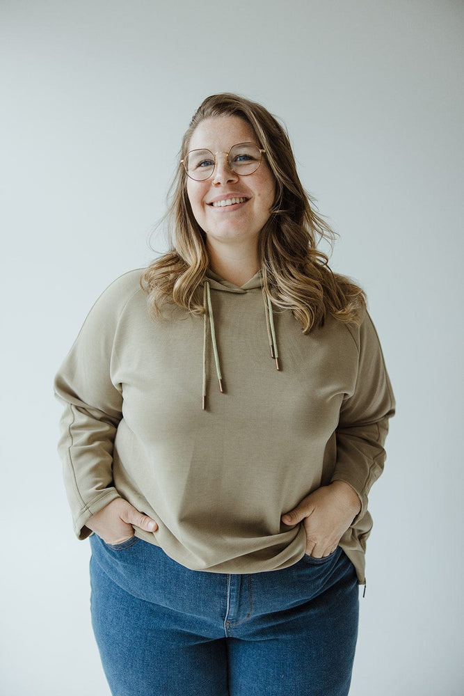 A person with long hair and glasses is standing against a plain background, wearing a Yesta Light as Air Double-String Hoodie in OM Green and blue jeans, epitomizing contemporary leisurewear.