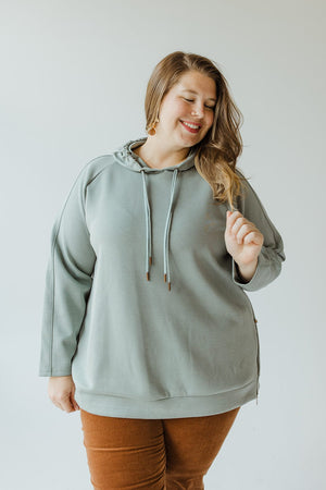 A person wearing a Yesta LIGHT AS AIR DOUBLE-STRING HOODIE IN QUIETUDE and brown pants stands against a plain background, smiling and looking slightly downward.