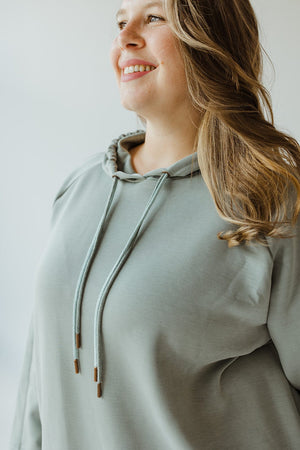 A woman is wearing Yesta's LIGHT AS AIR DOUBLE-STRING HOODIE IN QUIETUDE, smiling while looking slightly upwards. Her long, wavy hair rests over her shoulder, reflecting the luxurious comfort of contemporary leisurewear.