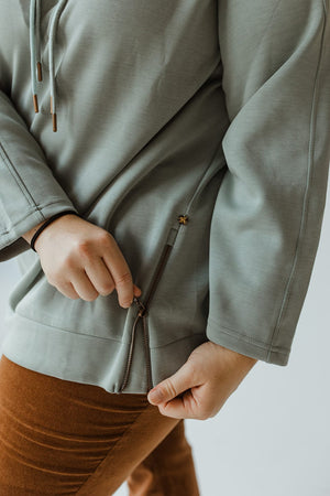 A person in the Yesta Light As Air Double-String Hoodie in Quietude, unzipping the side zipper near the hem, exudes luxurious comfort.