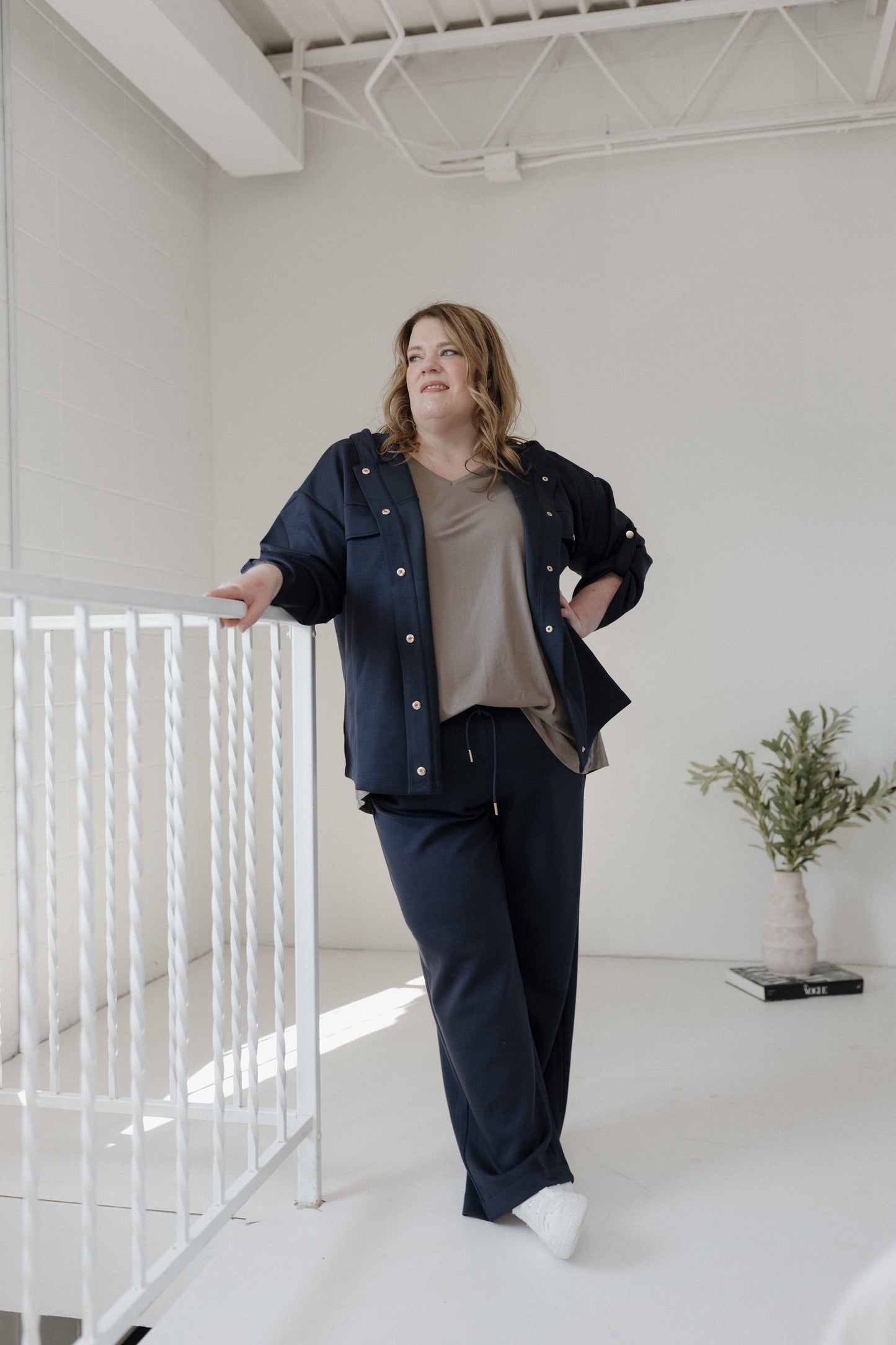 Woman in a navy outfit standing in a minimal white room with a plant.