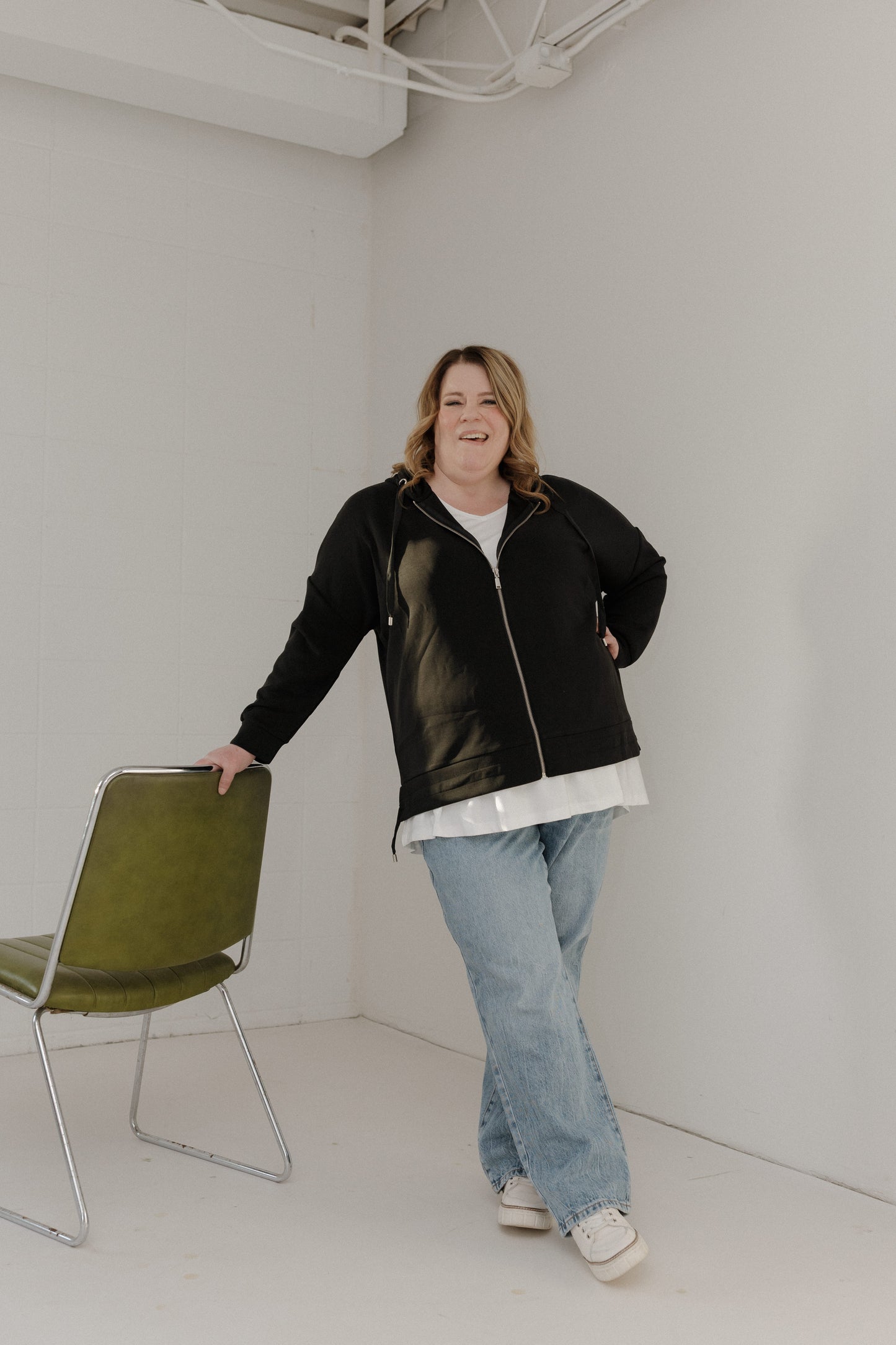 Person wearing a black jacket and blue jeans standing in a minimal white room with a green chair.