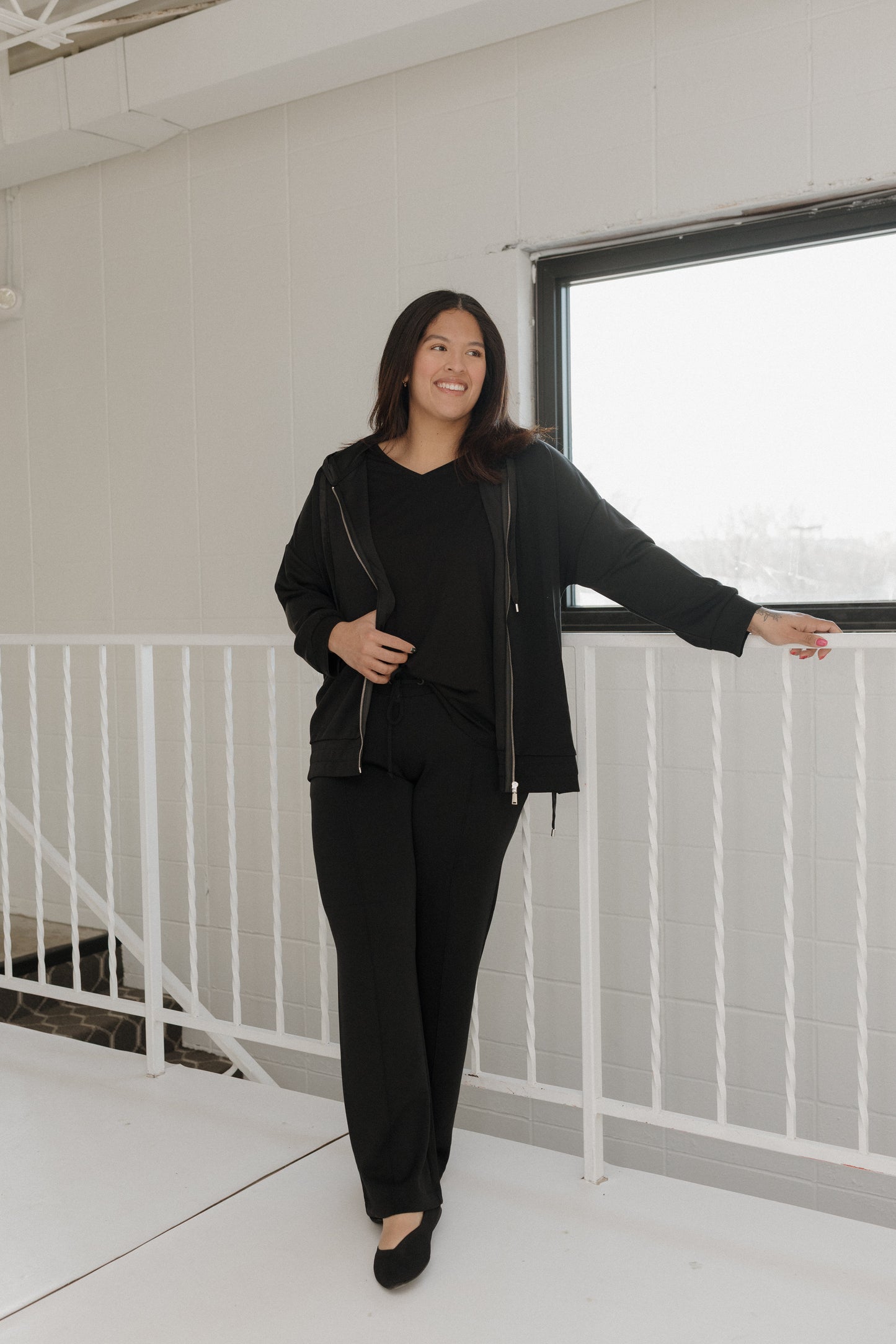 Woman in a black outfit standing in a modern indoor setting with a white railing and light-colored floor.