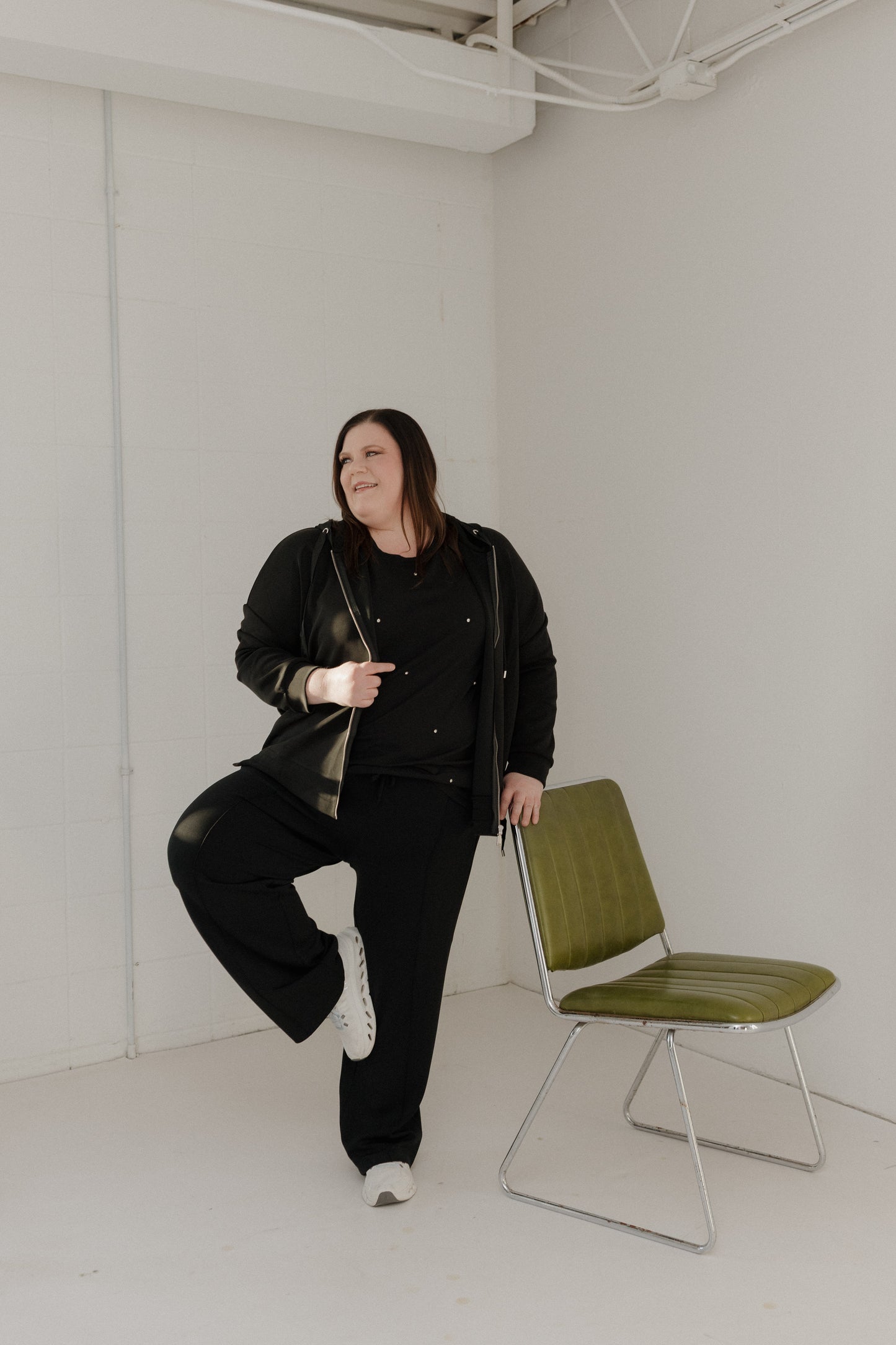 Woman in black outfit posing in a minimal white room with a green chair.