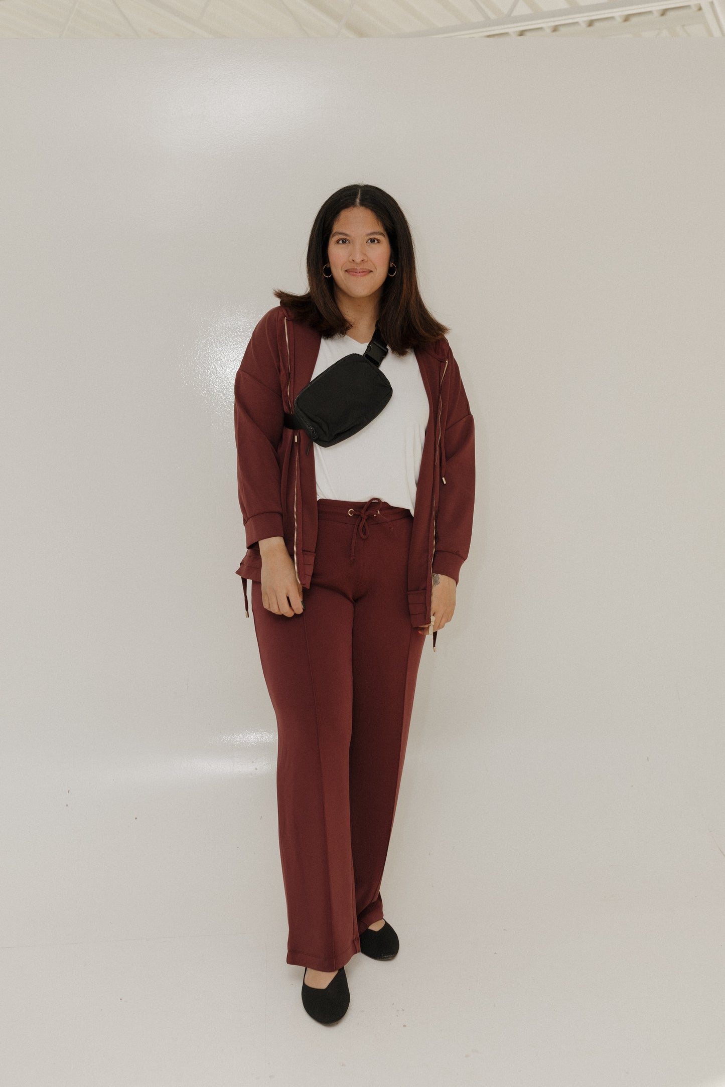 Woman wearing a maroon jacket and pants with a white shirt and black bag against a plain background
