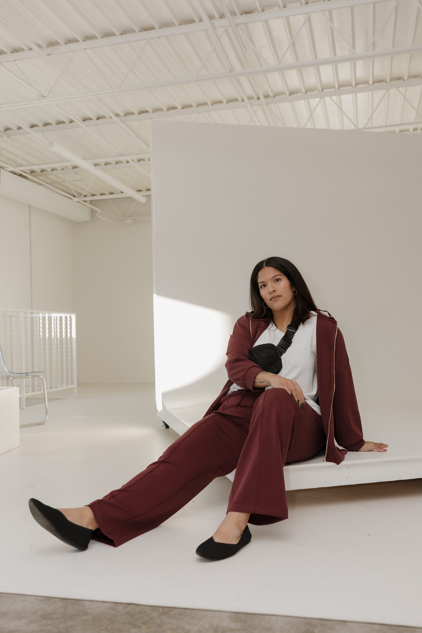 Woman in a maroon suit sitting on a white bench in a minimalistic room.