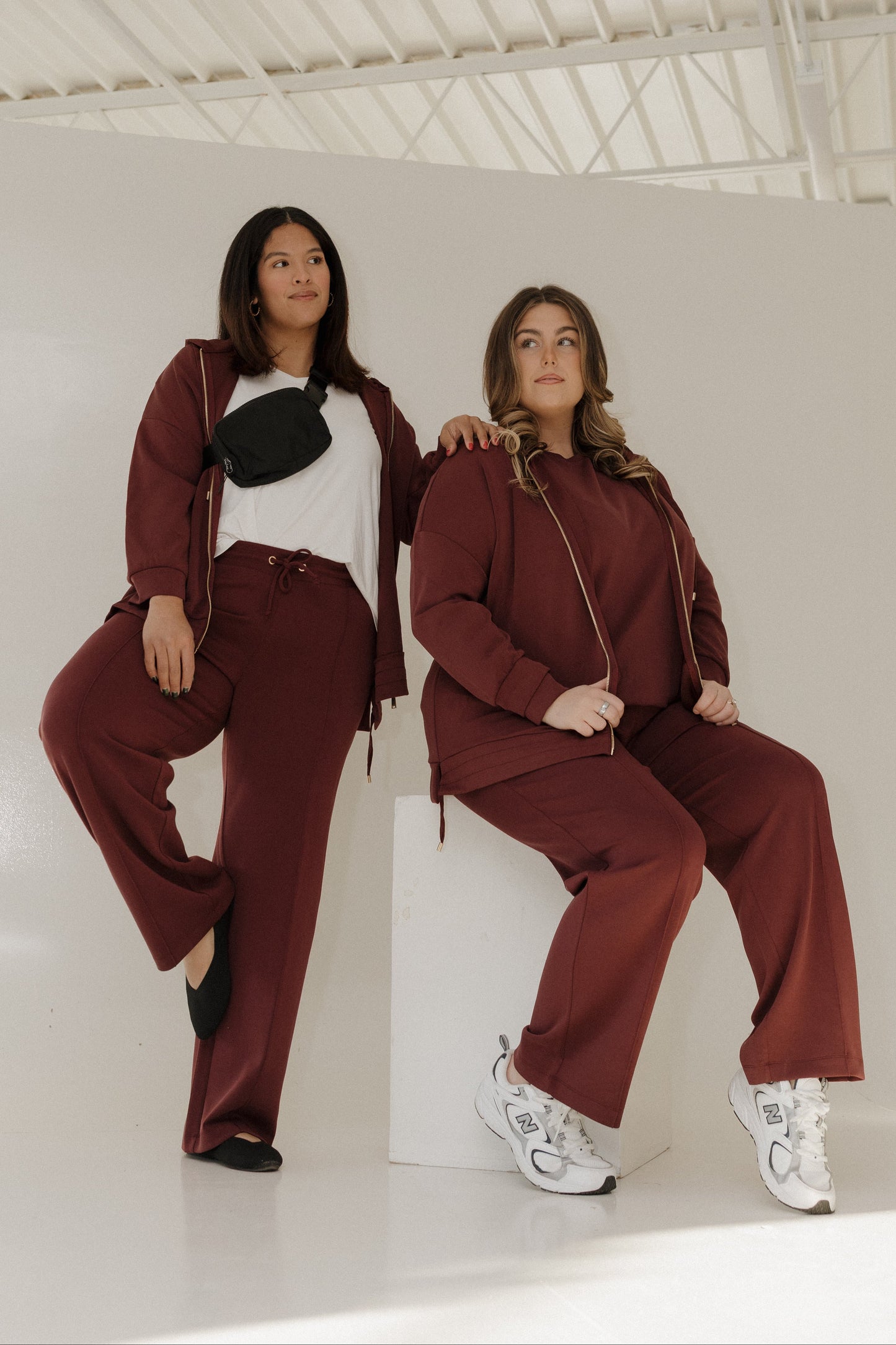 Two women in matching maroon tracksuits sitting on a white floor.