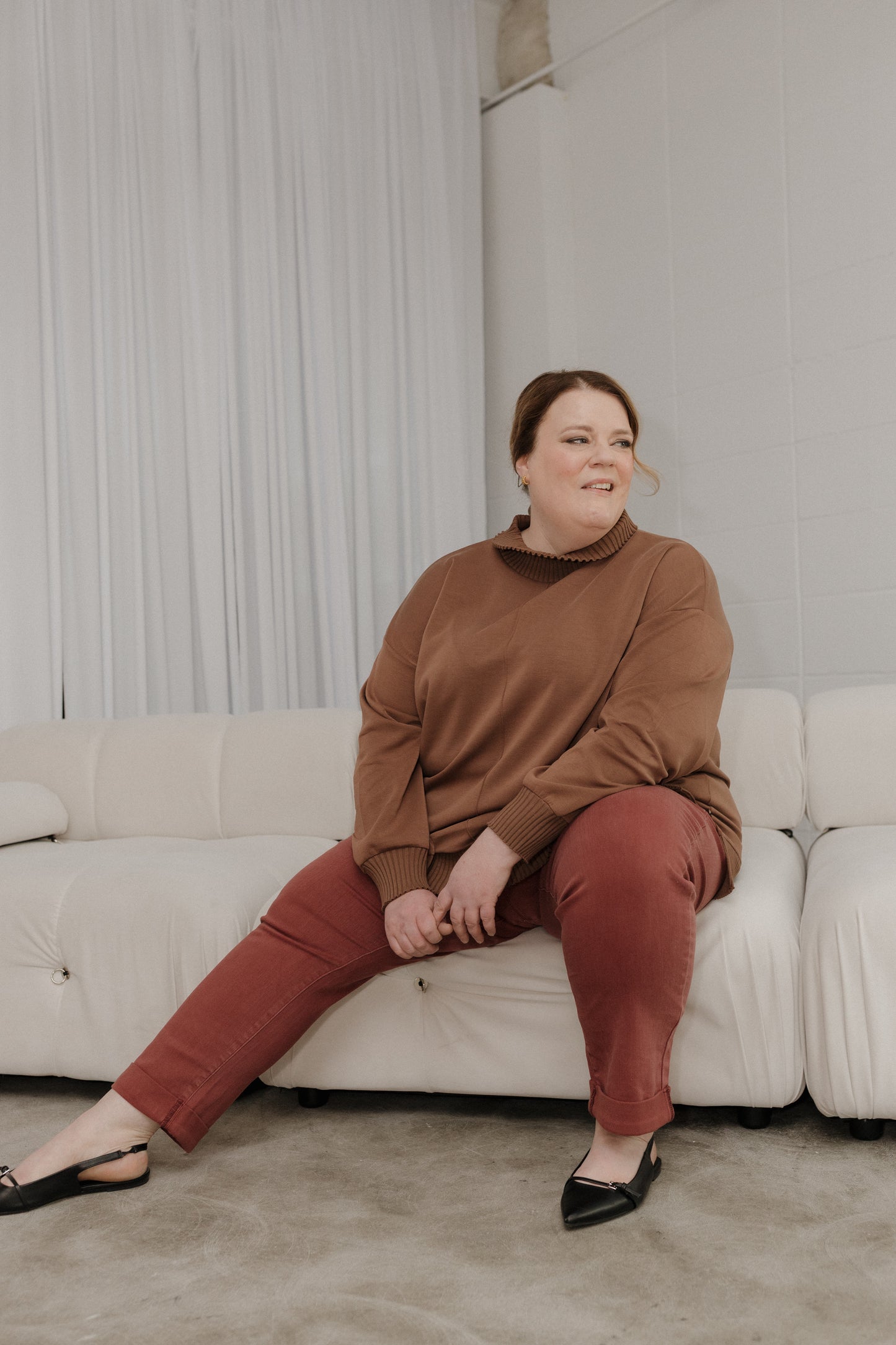Woman sitting on a white couch wearing a brown sweater and red pants in a neutral-colored room.