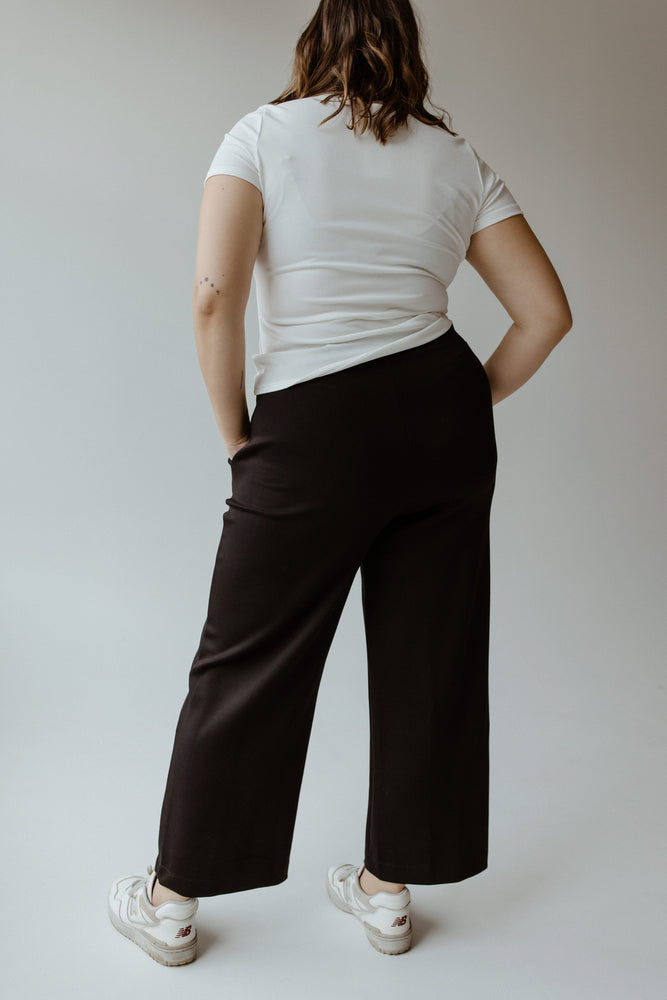 A person in a white shirt and Yesta's LIGHT AS AIR PLEATED WIDE LEG PANTS with an elastic waistband stands with hands in pockets, facing away.