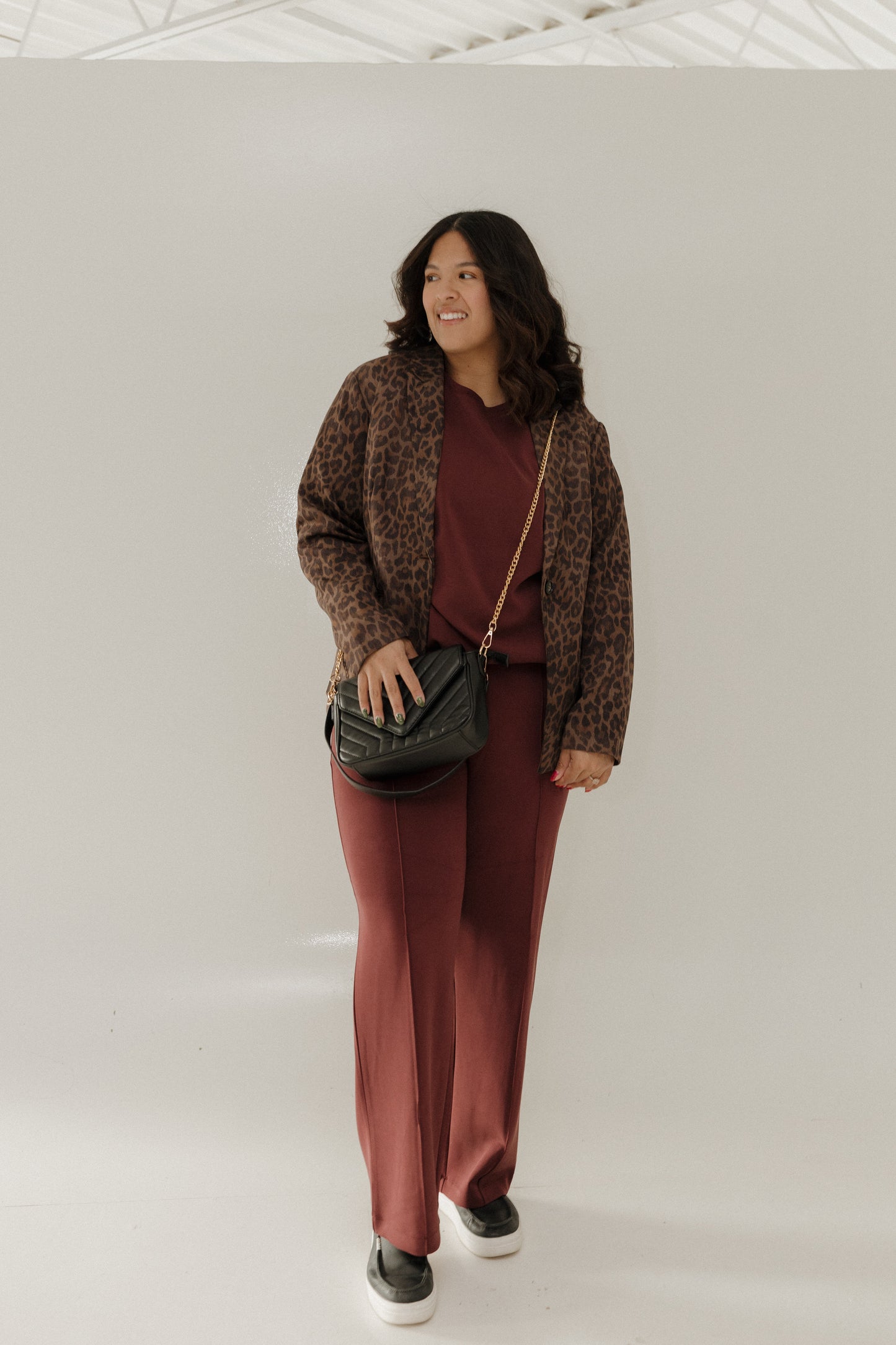 Woman wearing a brown patterned cardigan, burgundy top, and pants with a black handbag against a white background.