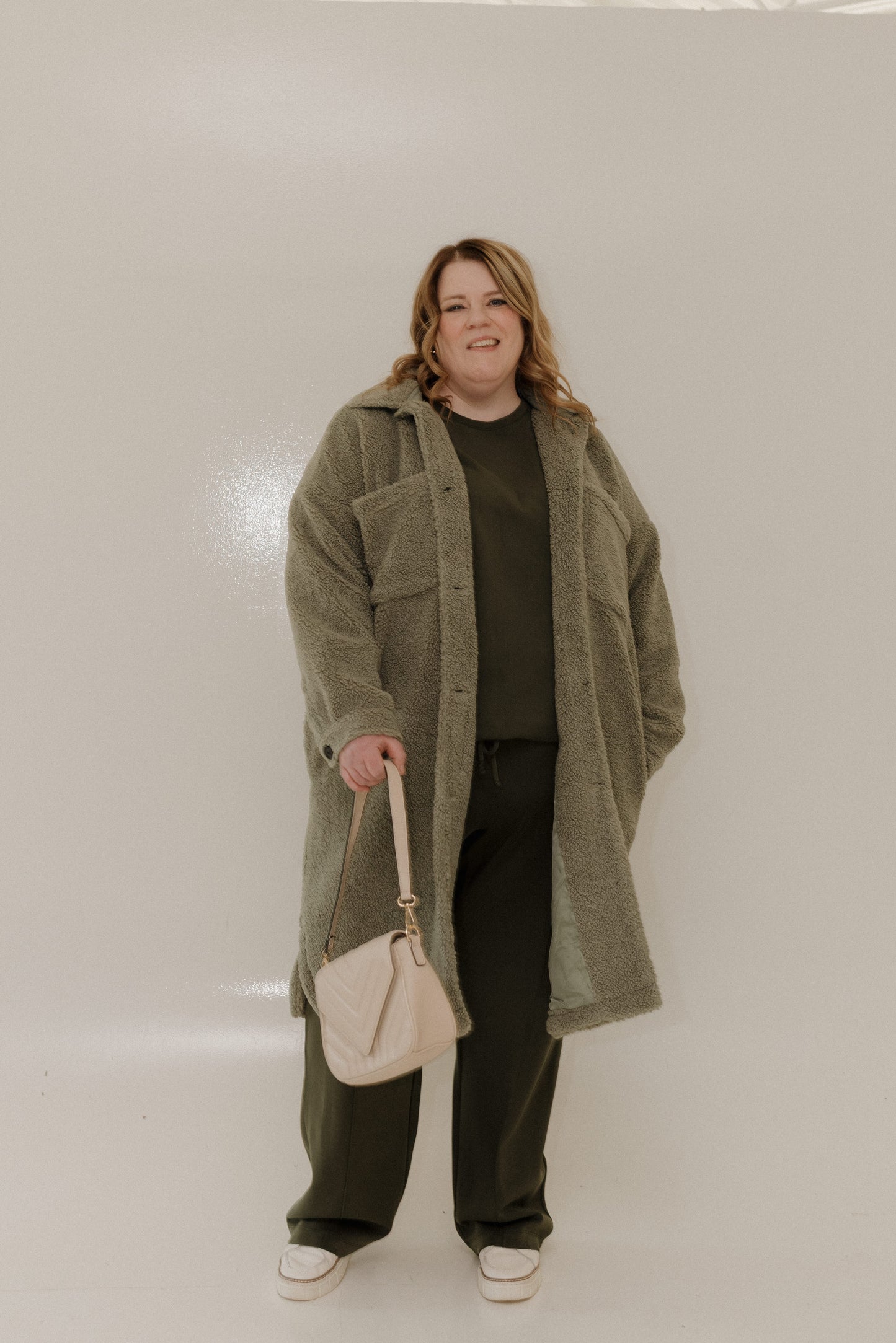 Person wearing a long olive green coat holding a beige handbag against a plain background