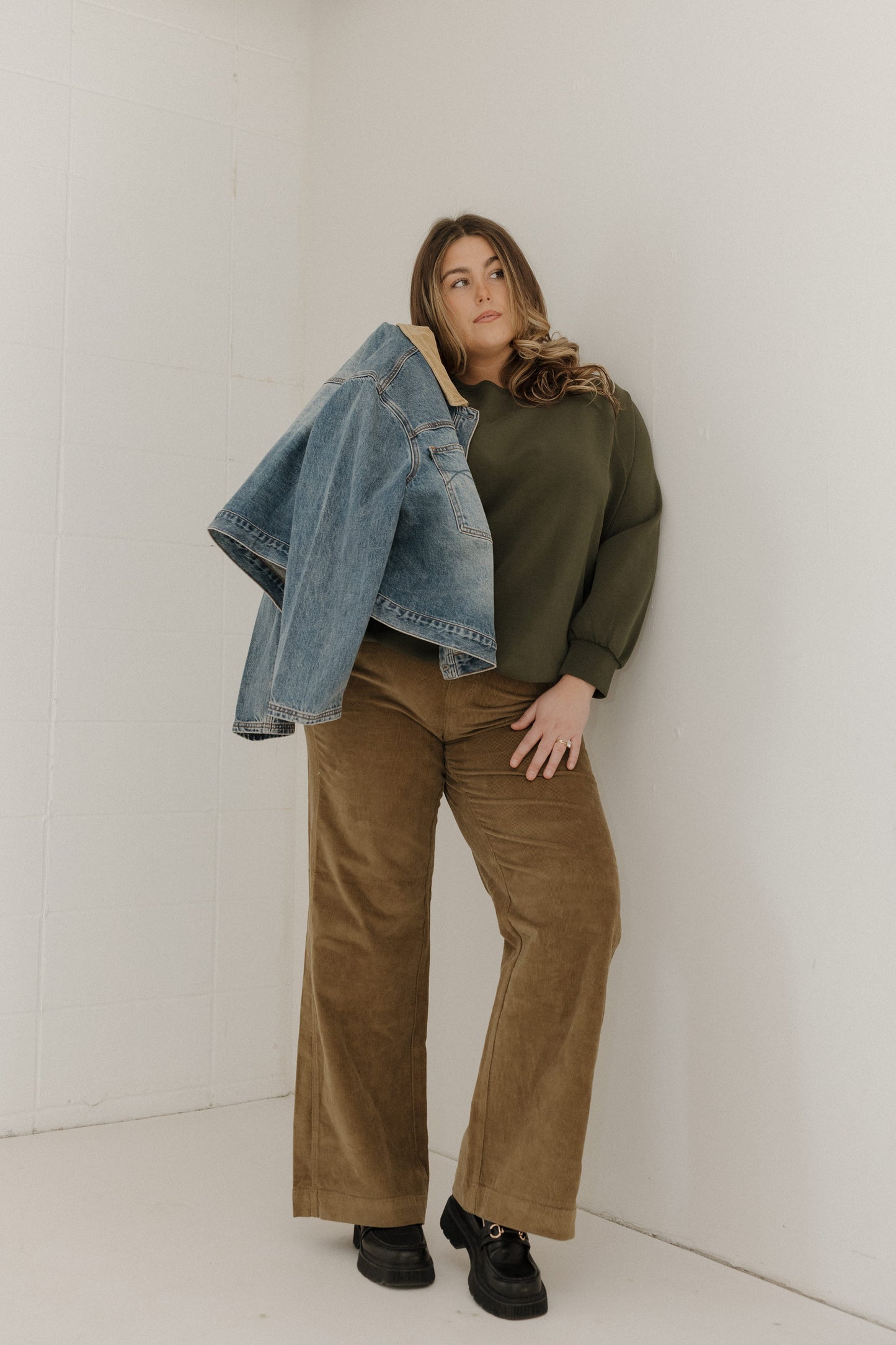 Woman wearing a green sweater, brown pants, and a denim jacket against a white wall.