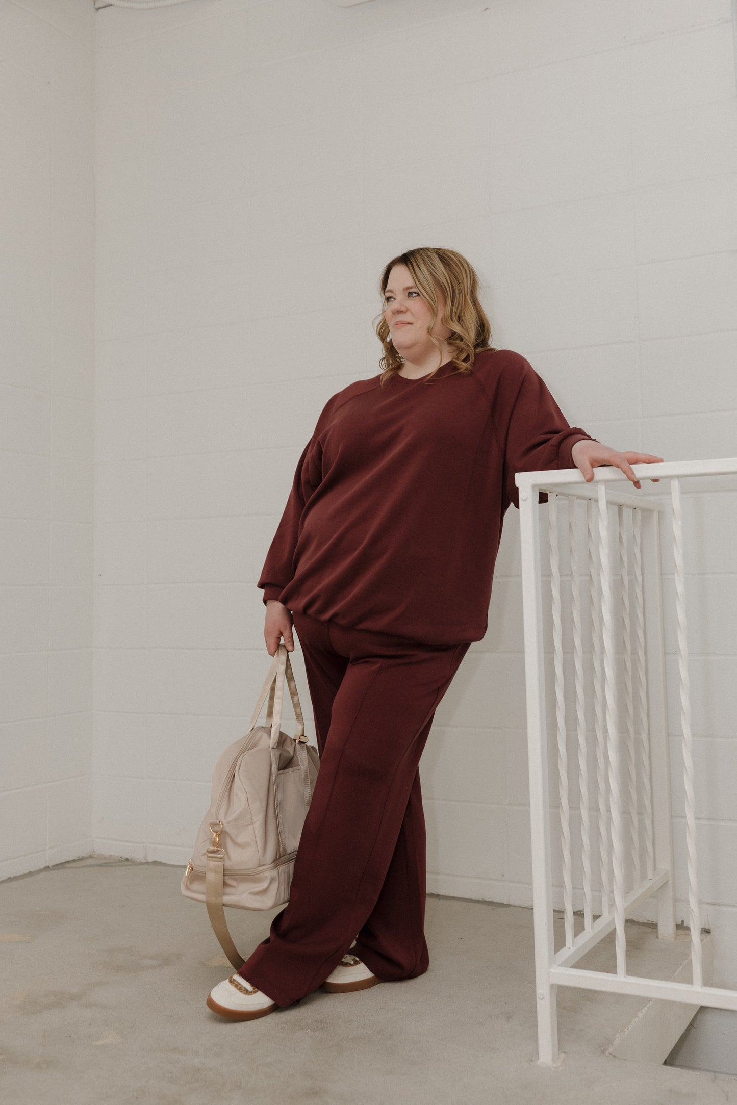 Woman in burgundy outfit holding a beige bag against a white wall.