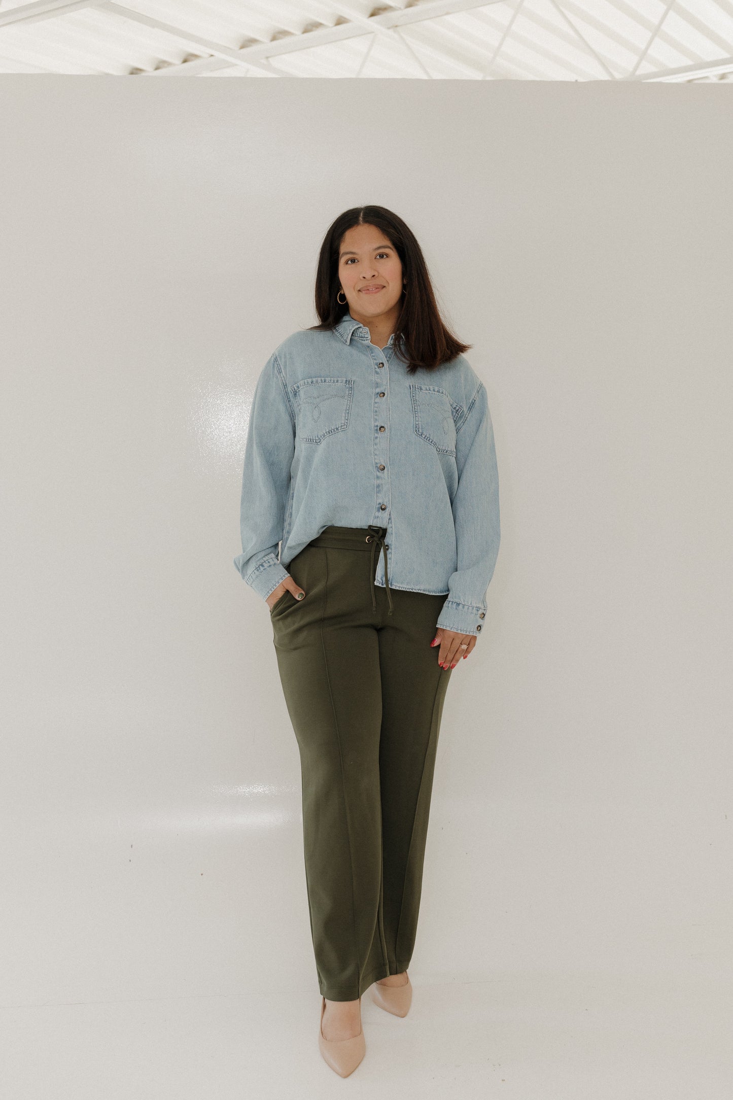 Woman wearing a light blue denim shirt and olive green pants against a white background