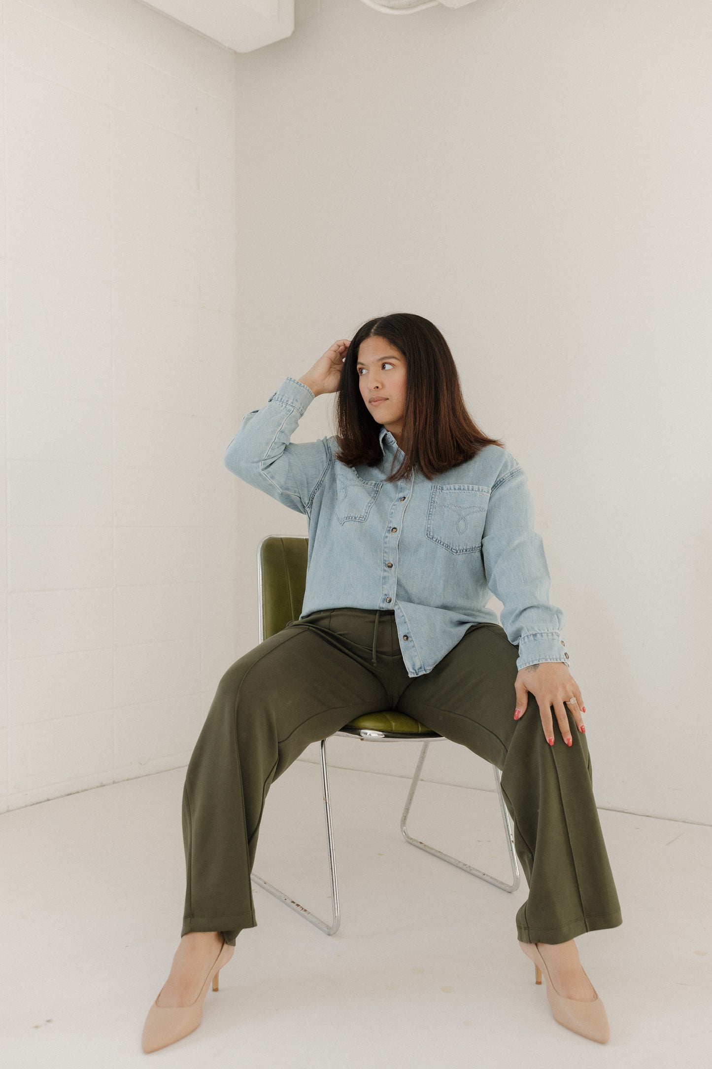Woman sitting on a chair wearing a light blue denim shirt and olive green pants in a minimal white room.