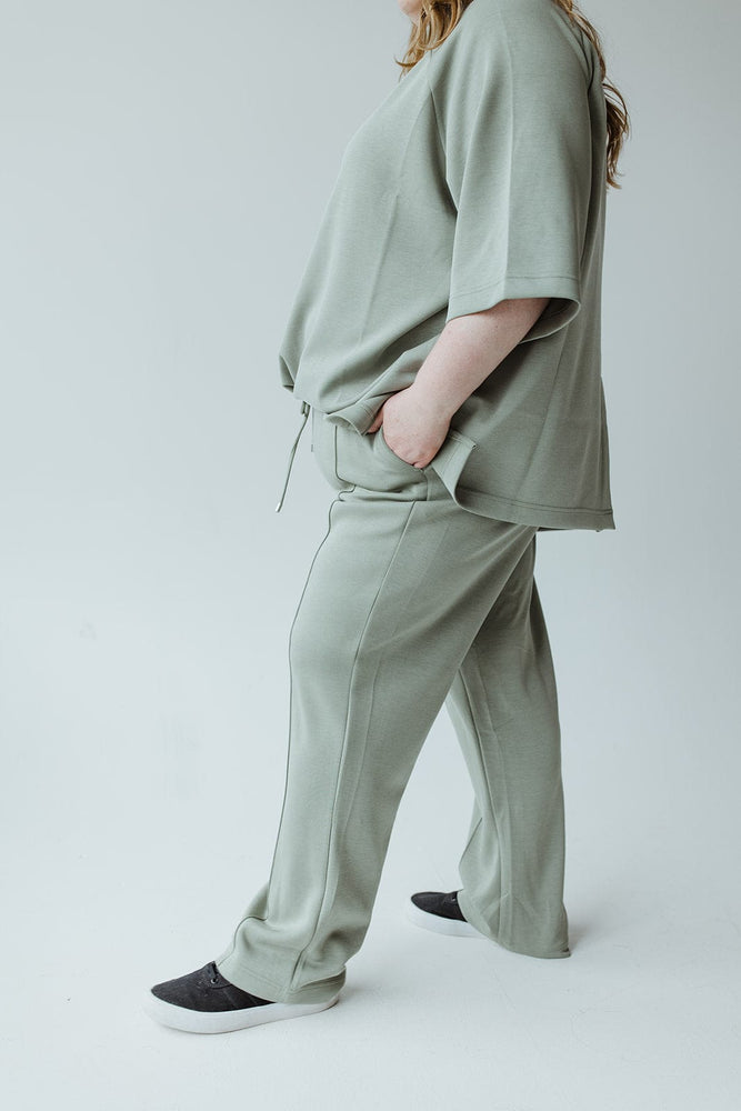 A person is wearing the LIGHT AS AIR TROUSER IN ECHEVERRIA by Yesta, a loose-fitting, matching green top and pants set featuring an elastic waistband and pockets. They are also wearing black shoes. Only part of their body is visible in the image.