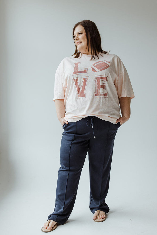 A woman with long hair stands against a plain background, wearing a pink "LOVE" t-shirt with a football image, Yesta's LIGHT AS AIR TROUSER IN MIDNIGHT featuring an elastic waistband, and flip flops. Her hands are in her pockets, and she appears relaxed.