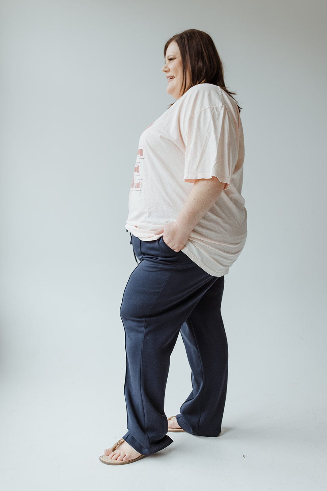 A person wearing a light pink shirt and the Yesta LIGHT AS AIR TROUSER IN MIDNIGHT stands in a relaxed pose, hands in pockets, with light sandals and a neutral backdrop.