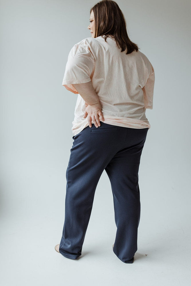 A person wearing a light-colored t-shirt and the Yesta LIGHT AS AIR TROUSER IN MIDNIGHT is standing with their back to the camera, slightly turned to the left, with one hand on their lower back.