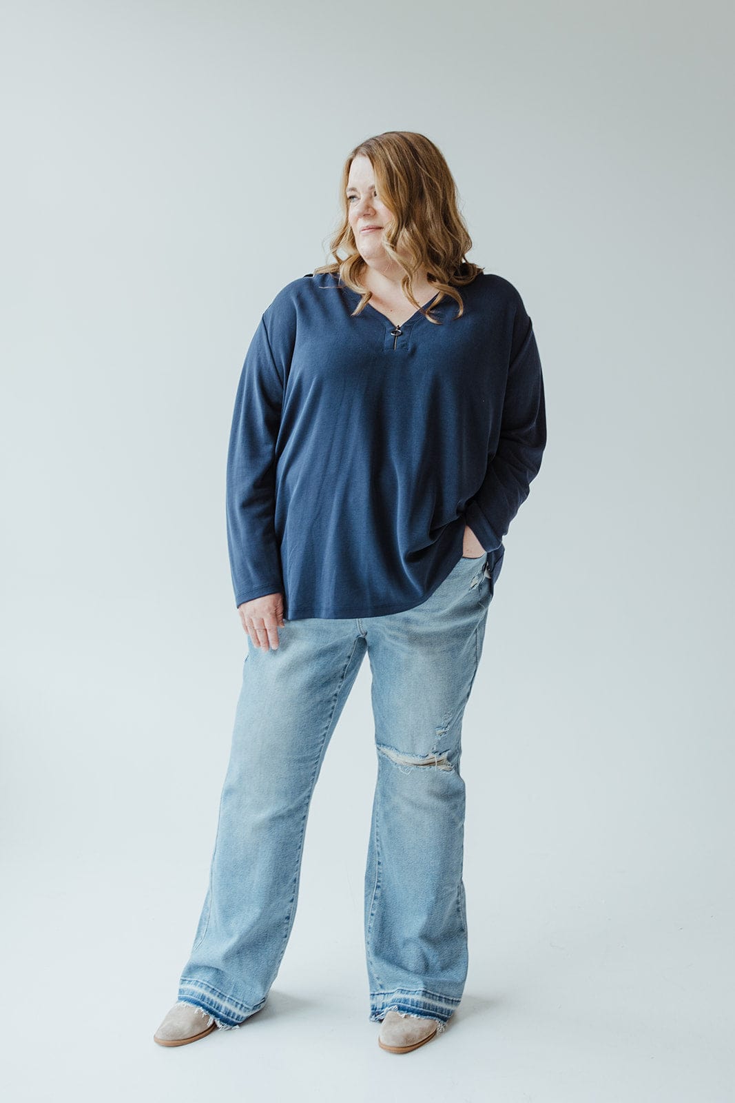 A woman with light brown hair is standing against a plain backdrop. She is wearing the navy blue "Light Texture Tunic with Zipper" by Yesta, paired with light blue ripped jeans and beige shoes. One hand rests in her pocket.