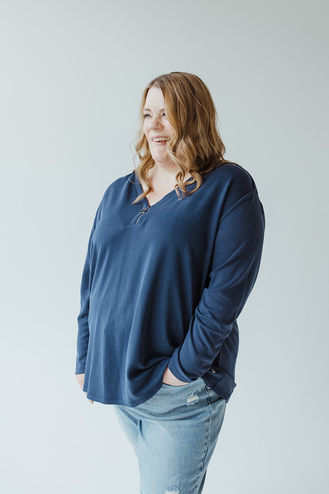 A person with long wavy hair is standing against a plain light background, wearing the Yesta LIGHT TEXTURE TUNIC WITH ZIPPER in navy blue along with light blue jeans.