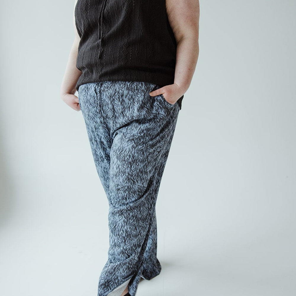 A person wearing a sleeveless black top, blue linear-pattern woven pants with an elastic waistband, and black sandals stands with hands in pockets against a plain background.