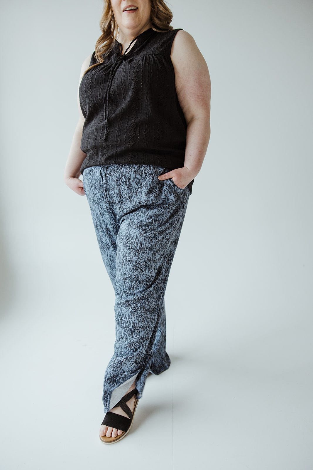 A person wearing a sleeveless black top, blue linear-pattern woven pants with an elastic waistband, and black sandals stands with hands in pockets against a plain background.