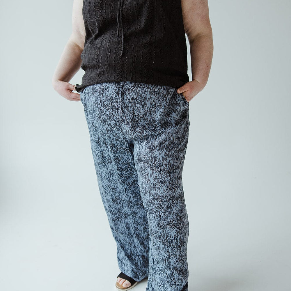 A person wearing a sleeveless black top and patterned blue pants with an elastic waistband stands with hands in pockets against a plain background.