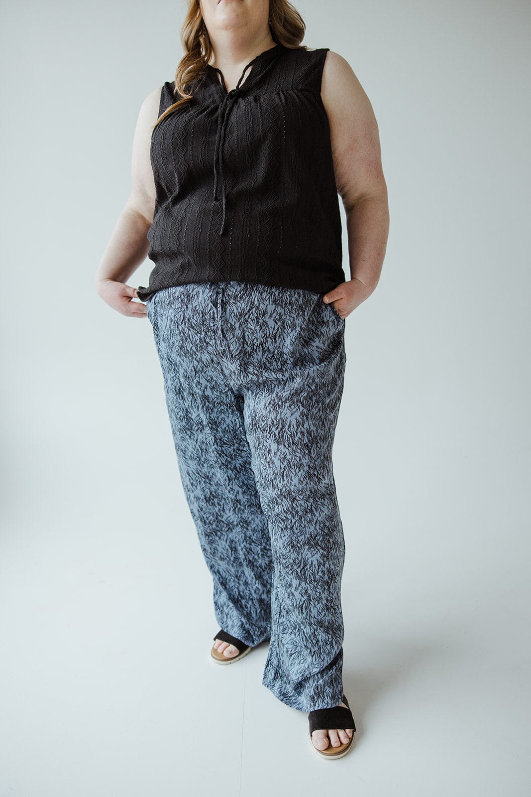 A person wearing a sleeveless black top and patterned blue pants with an elastic waistband stands with hands in pockets against a plain background.