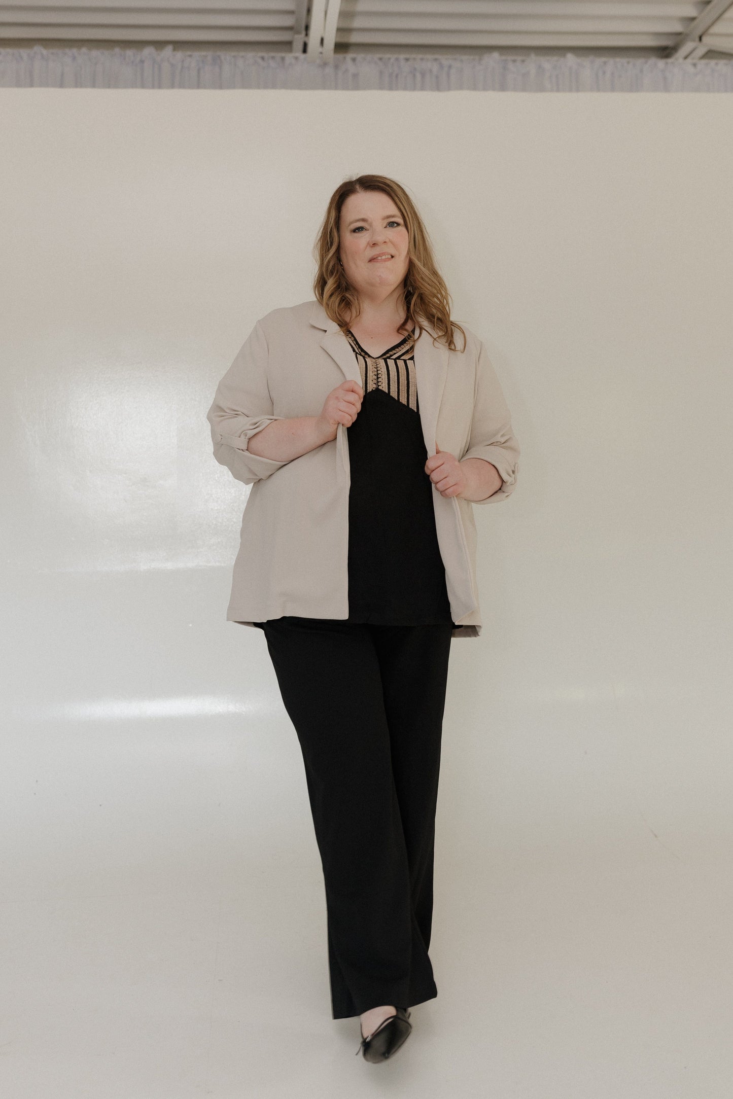 Woman wearing a beige jacket over a black top and pants against a white wall.
