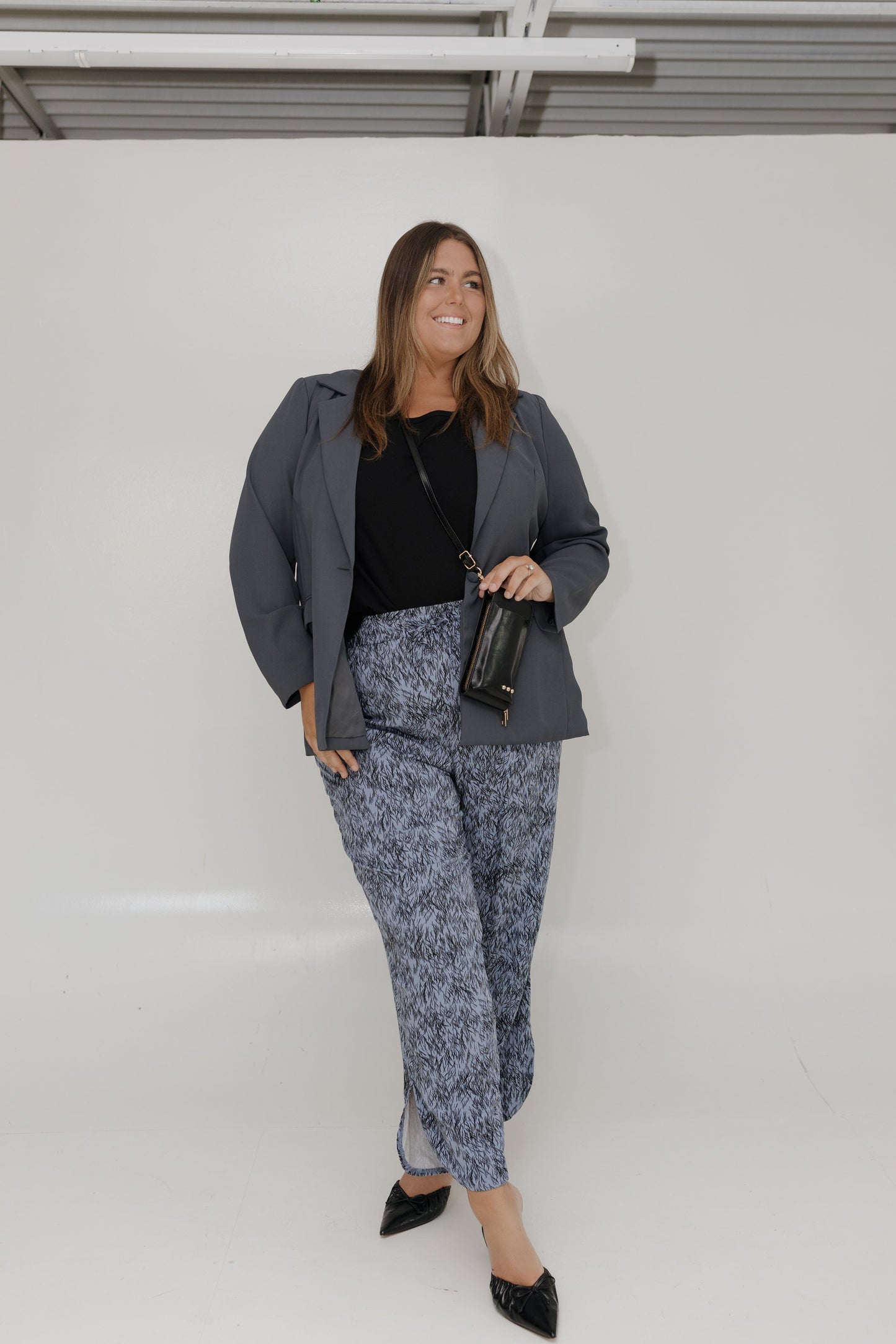 Woman in a gray blazer and patterned pants standing against a white wall.