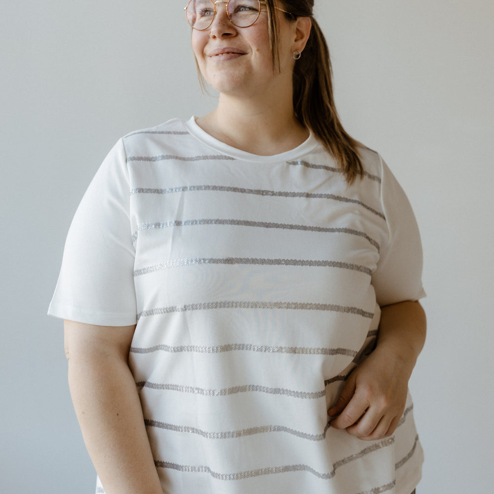 A person with glasses and a ponytail, wearing a Sequin Stripe Tee made of soft fabric and gray pants, stands against a plain background, looking slightly to the side.